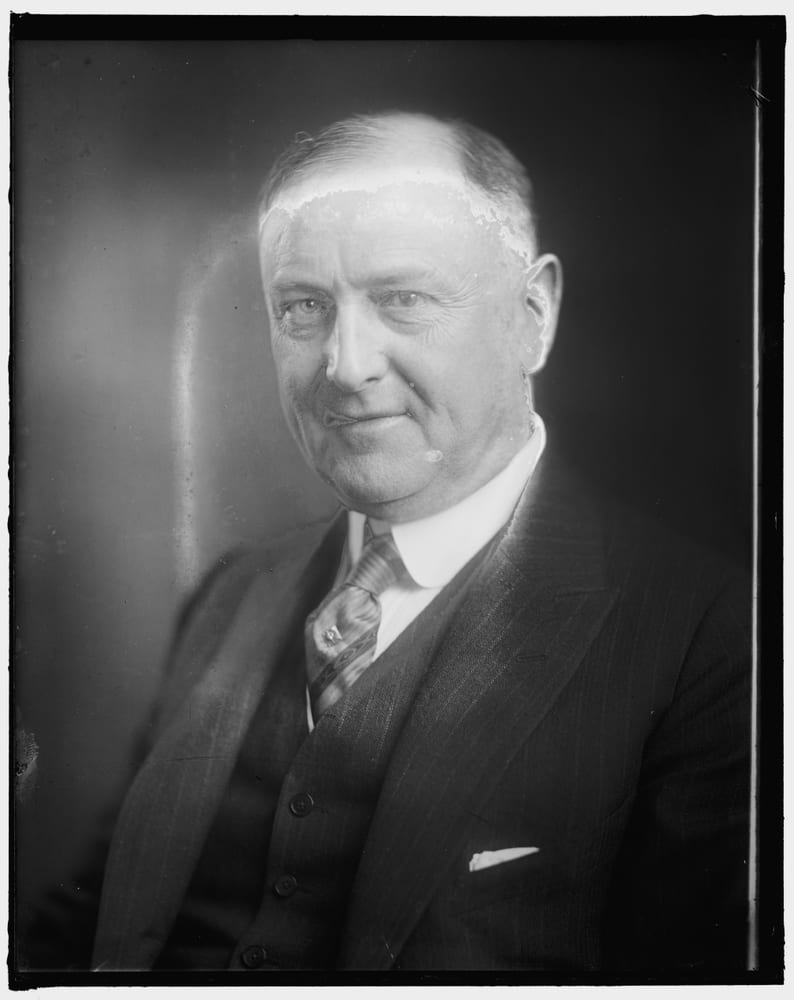 A glass-negative portrait of an older man in a suit, with emulsion flaking along the hairline and a pale streak at the left.