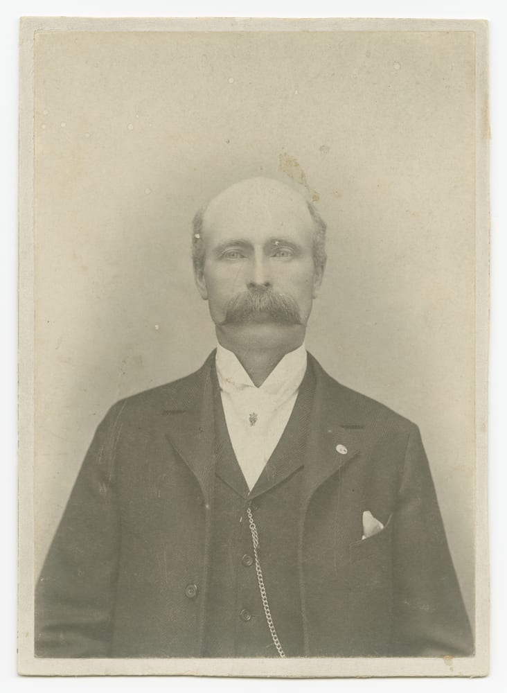 A faded cabinet card of a man with a mustache, in a dark suit, the print soft and low in contrast.